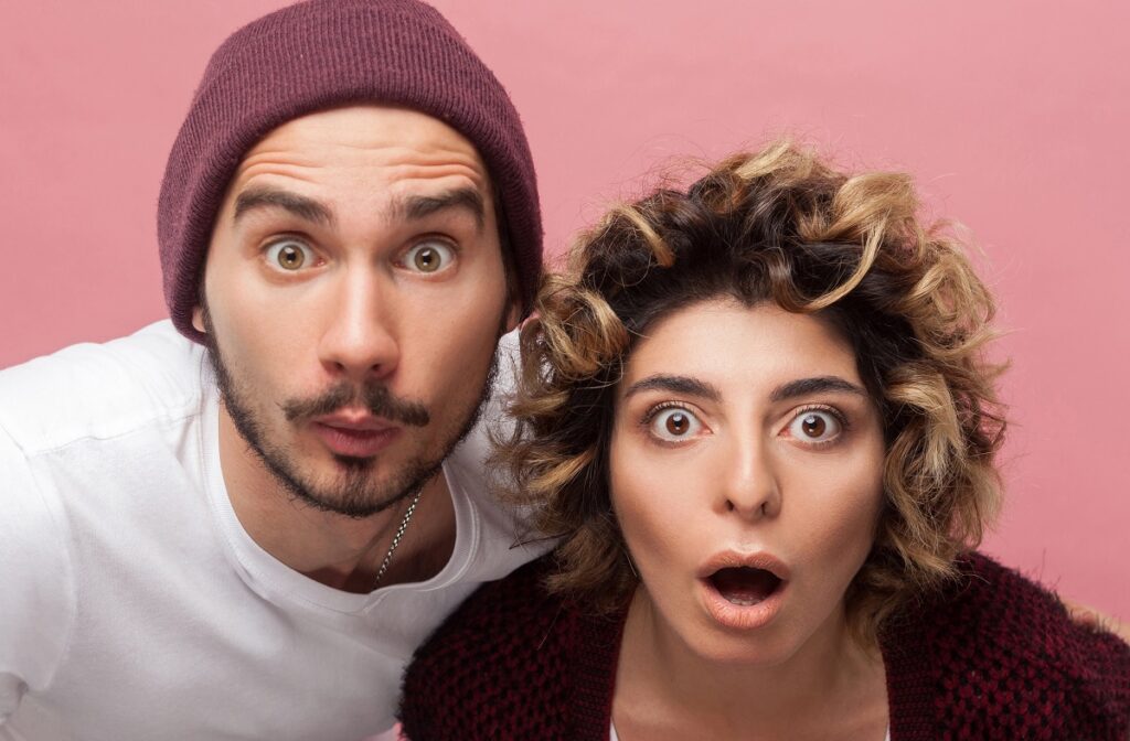 Closeup portrait of shocked funny couple of friends in casual style standing with unbeliveable face and looking at camera with big eyes and opened mouth. Isolated,indoor, studio shot, pink background