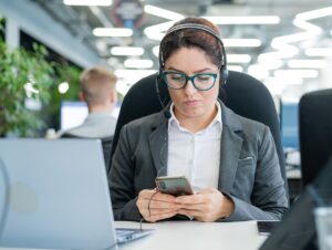 Business woman dressed in a headset is bored and uses a smartphone while sitting at a desk. Female manager is distracted from work by phone