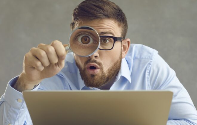 Portrait of funny handsome nerdy young business man in glasses sitting at office desk with laptop computer, holding magnifying glass and looking at something with big eye and surprised face expression