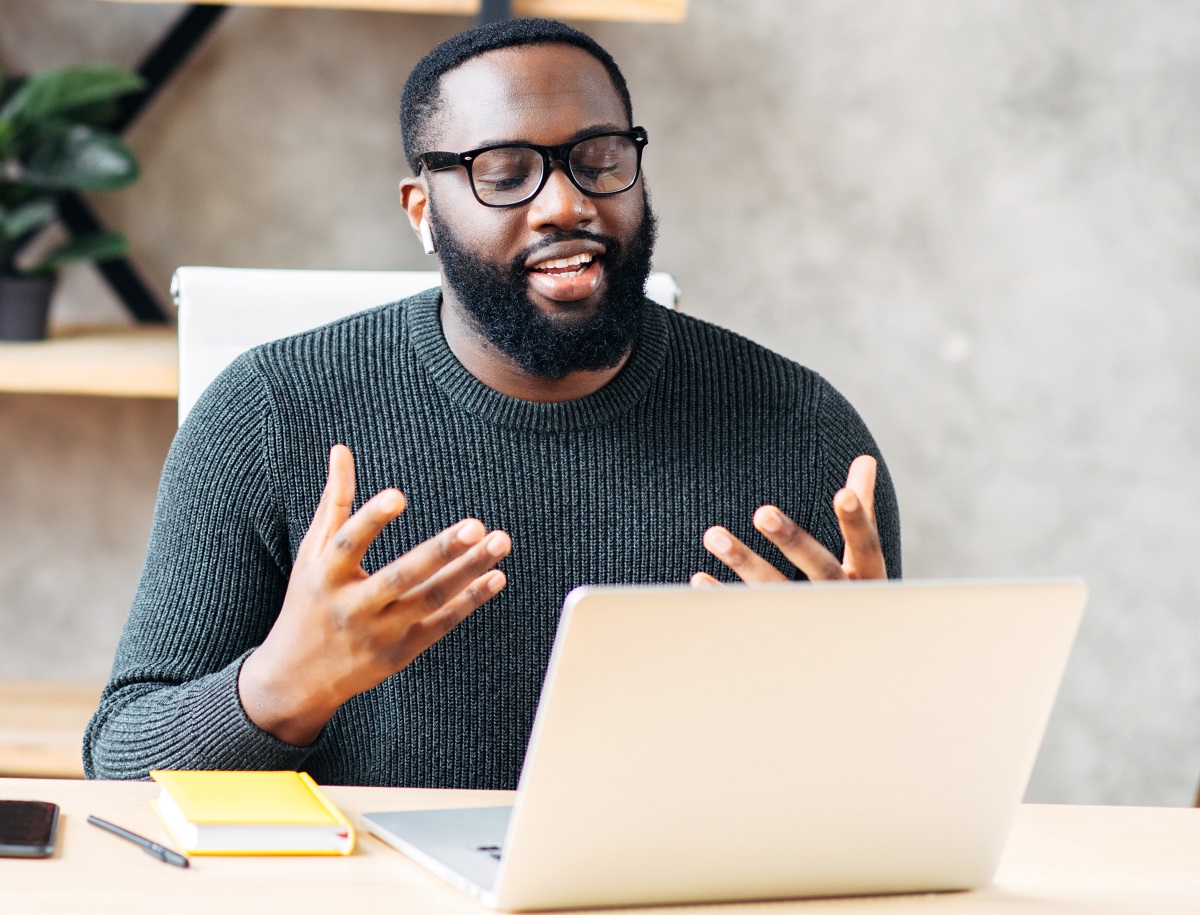 Video call. Attractive African-American guy is using laptop for video communication, a guy in glasses sits at the table in modern office, looking into webcam and talking online